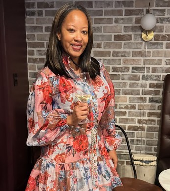 Qiana Taylor holding a glass of white wine, smiling warmly in a floral dress against an exposed-brick wall