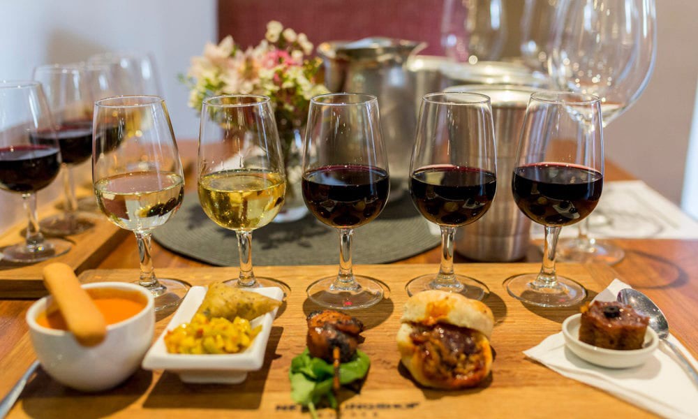 A tasting flight of red and white wines, each glass paired with a small plate on a wooden board