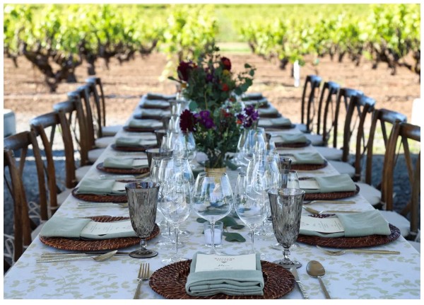 A long dinner table set with glasses and florals between vineyard rows