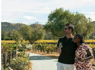A couple strolls a sunlit vineyard path beneath olive trees on an estate tour